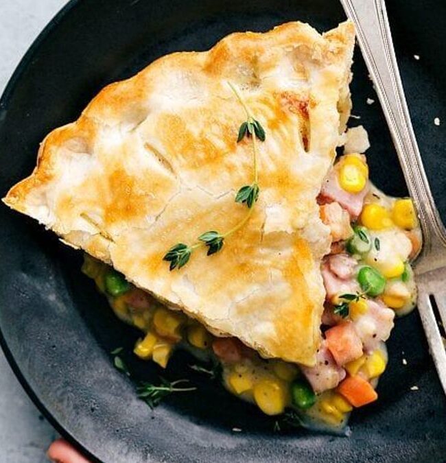 A slice of vegetable pot pie on a black plate, garnished with fresh herbs. The pie has a golden, flaky crust and is filled with peas, corn, and carrots. A fork rests beside the slice.