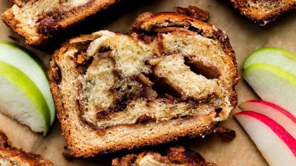 Close-up of a slice of cinnamon babka with visible swirls of cinnamon and sugar in the bread, offering a modern twist to old-fashioned desserts. Next to it are slices of green and red apples on a parchment paper background. The babka boasts a golden-brown crust.