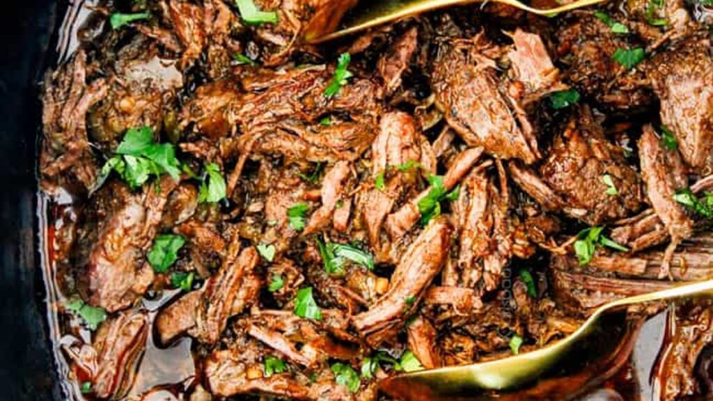 Shredded beef is garnished with chopped fresh herbs. The dish is moist and tender, with visible rich juices. Two metal serving spoons are placed among the meat.