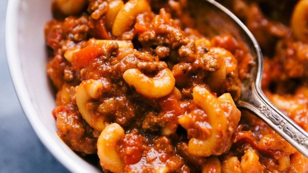 A close-up of a bowl of goulash featuring macaroni pasta, ground meat, and a rich tomato sauce. A spoon is partially submerged on the right side, showcasing the dish's hearty texture. The dish appears warm and savory.