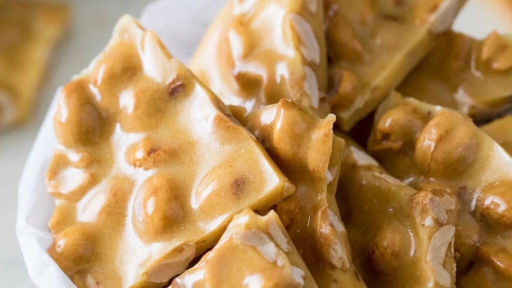 Close-up of peanut brittle pieces, showcasing a glossy, golden-brown caramelized surface with embedded peanuts. The jagged, uneven shapes are piled together, highlighting the texture and shine of the treat.