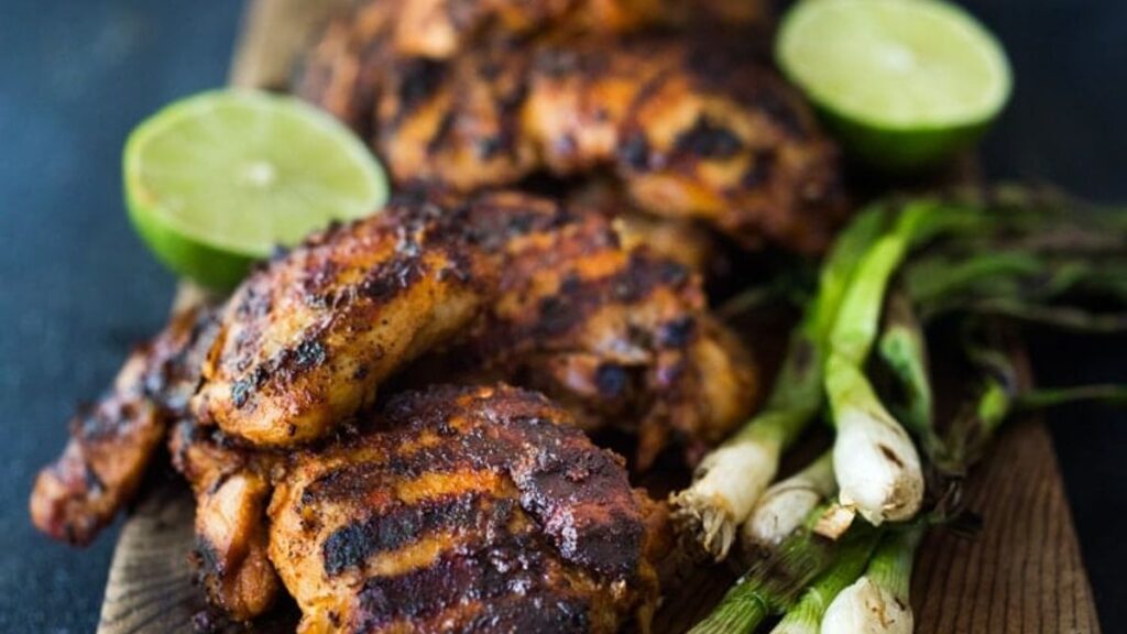 Grilled chicken pieces with char marks are presented on a wooden board, accompanied by fresh green onions and halved limes. The dish is set on a dark surface.