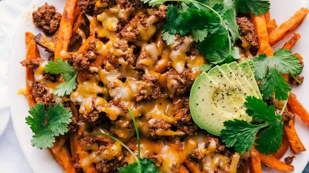 A close-up of sweet potato fries topped with melted cheese, seasoned ground meat, sliced avocado, and fresh cilantro on a white plate.