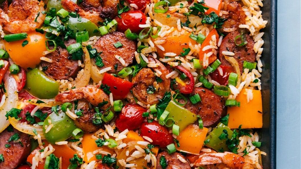 A vibrant one-pan dish of seasoned rice with shrimp, sliced sausage, and diced bell peppers, perfect for weeknight dinners. Red onions and cherry tomatoes add flavor, while chopped green onions and fresh herbs offer a colorful and appetizing presentation.