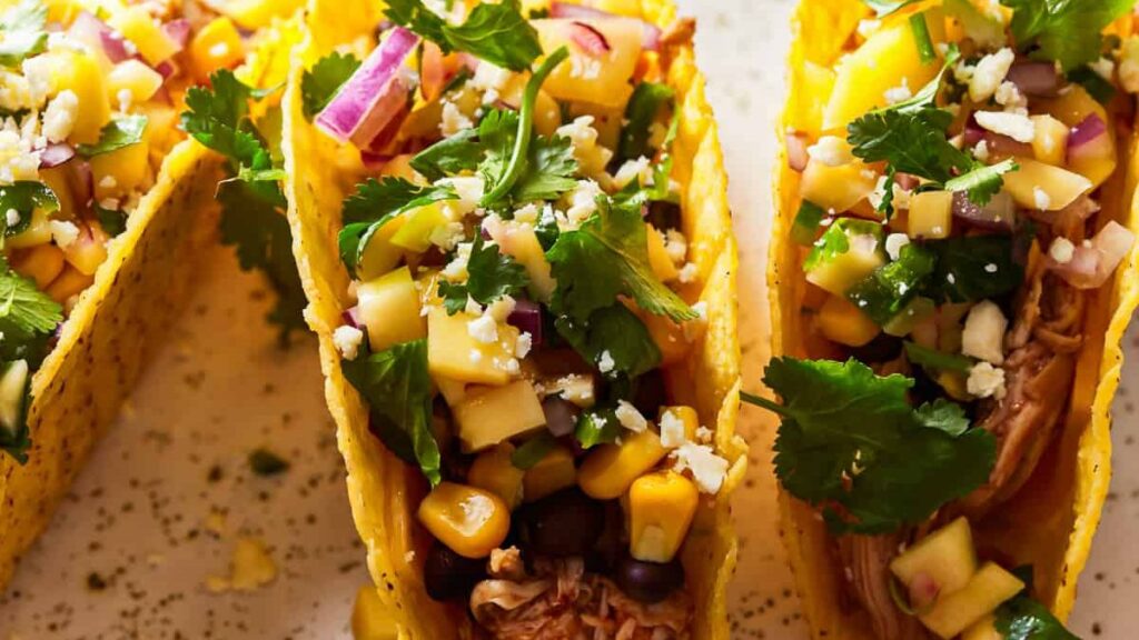 Close-up of three tacos filled with shredded meat, corn, black beans, diced mango, chopped red onions, cilantro, and crumbled cheese. Prepared using a slow cooker to make life easier, they're in yellow corn shells and garnished with fresh cilantro leaves.