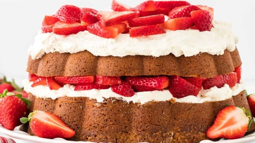 A layered strawberry shortcake with whipped cream between two layers of cake, topped with sliced fresh strawberries. Whole strawberries are scattered around the cake on a white plate.