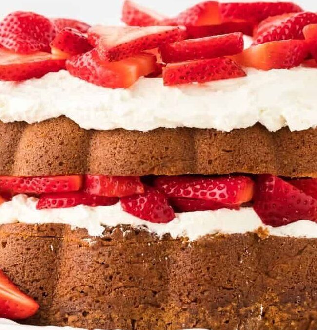 A layered strawberry shortcake with whipped cream between two layers of cake, topped with sliced fresh strawberries. Whole strawberries are scattered around the cake on a white plate.