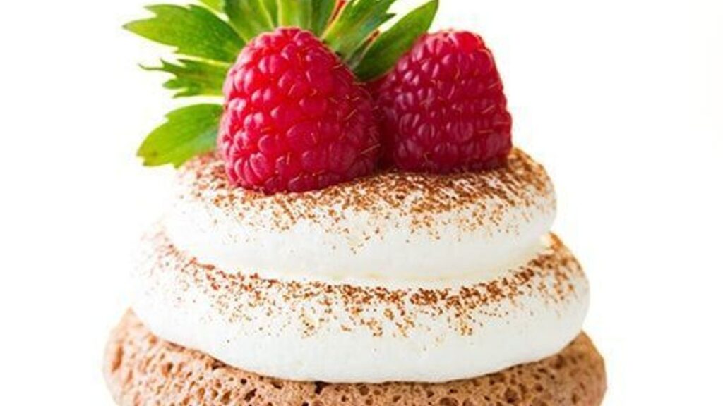 A dreamy dessert with a chocolate sponge base topped by two swirls of whipped cream, gently dusted with cocoa powder. Two fresh raspberries and a green mint leaf add a delightful garnish. The pristine white background completes the picture-perfect presentation.