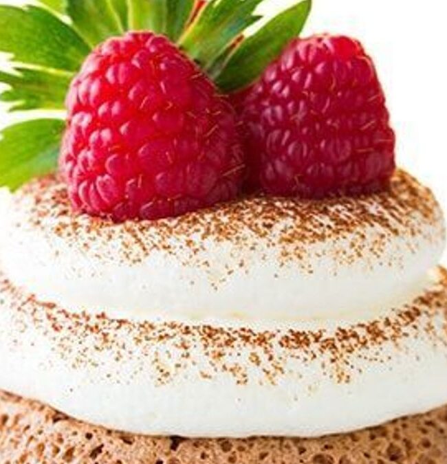 A dreamy dessert with a chocolate sponge base topped by two swirls of whipped cream, gently dusted with cocoa powder. Two fresh raspberries and a green mint leaf add a delightful garnish. The pristine white background completes the picture-perfect presentation.