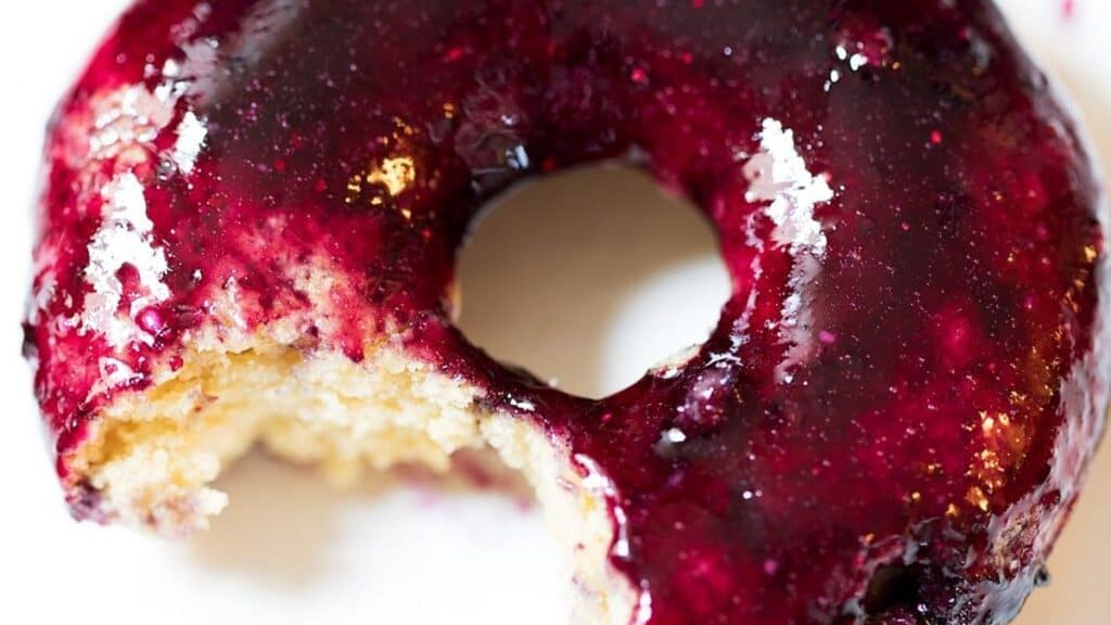 A close-up of a glazed donut with a deep purple berry topping. A small bite has been taken from the donut, revealing its light, fluffy interior. The glaze appears glossy under the light.