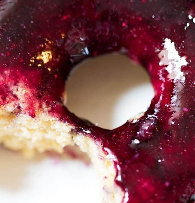 A close-up of a glazed donut with a deep purple berry topping. A small bite has been taken from the donut, revealing its light, fluffy interior. The glaze appears glossy under the light.