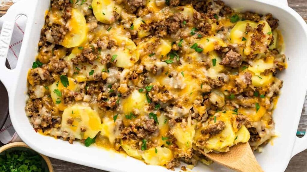 A casserole dish filled with layers of sliced potatoes, ground beef, and melted cheese, garnished with chopped parsley. This cozy casserole recipe is completed by a wooden spoon resting inside and a cloth napkin partially visible underneath the dish.