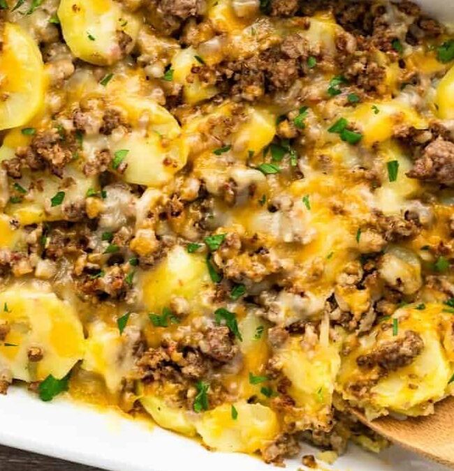 A casserole dish filled with layers of sliced potatoes, ground beef, and melted cheese, garnished with chopped parsley. This cozy casserole recipe is completed by a wooden spoon resting inside and a cloth napkin partially visible underneath the dish.