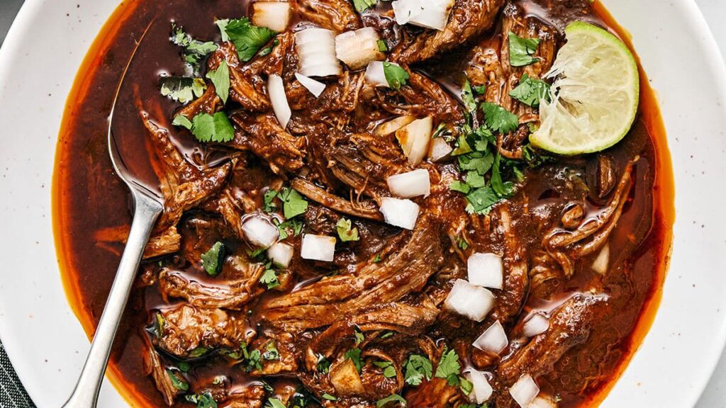 A white bowl filled with shredded beef birria stew in a rich, red-brown broth. Topped with diced onions and fresh cilantro, with a lime wedge on the side. A spoon is on the left side of the bowl.