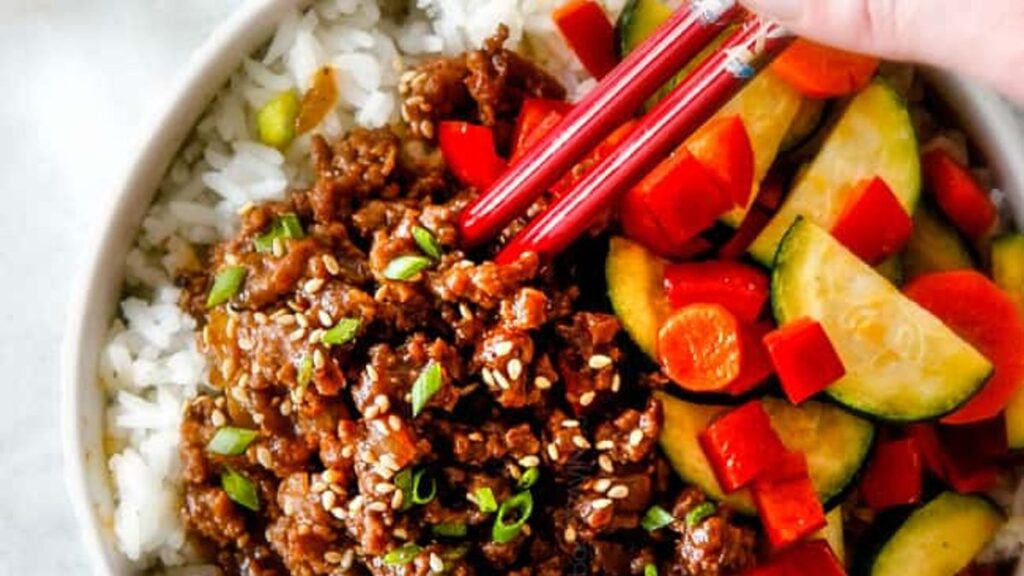 A bowl of rice topped with seasoned ground meat garnished with sesame seeds and chopped green onions. Sliced vegetables, including red bell peppers, zucchini, and carrots, are on the side. A hand is using red chopsticks to pick up some food.