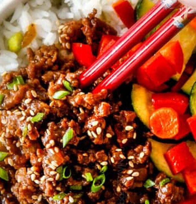 A bowl of rice topped with seasoned ground meat garnished with sesame seeds and chopped green onions. Sliced vegetables, including red bell peppers, zucchini, and carrots, are on the side. A hand is using red chopsticks to pick up some food.