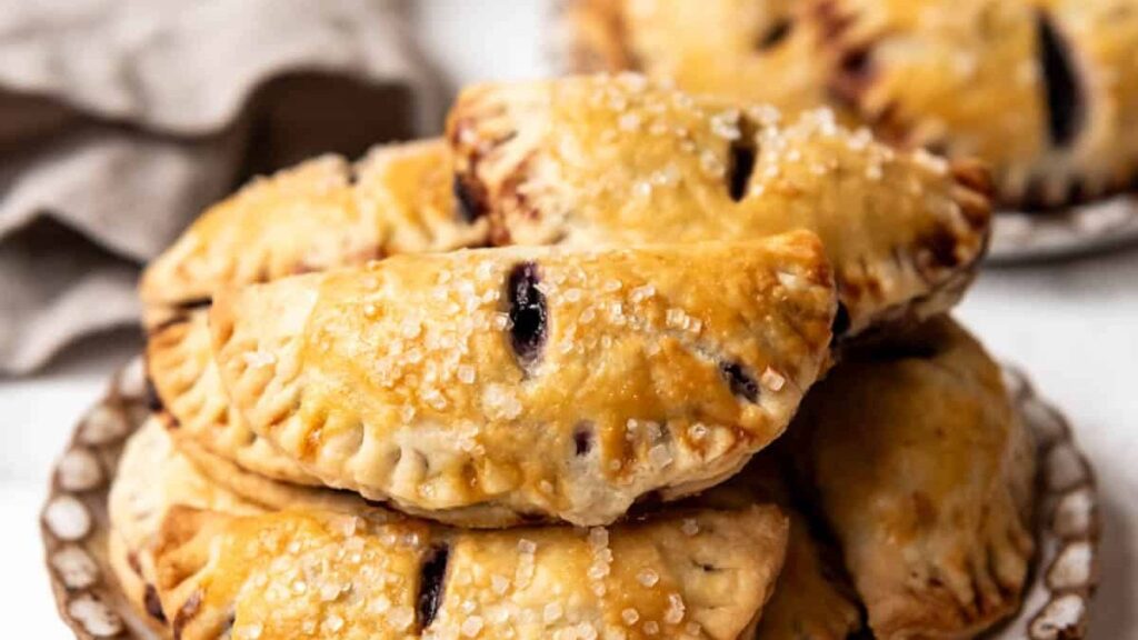 A plate full of indulgent golden-brown hand pies. The flaky, sugar-dusted crusts have small slits on top, adding charm to the baked delights neatly stacked. A beige cloth is partially visible in the background, enhancing their irresistible allure.